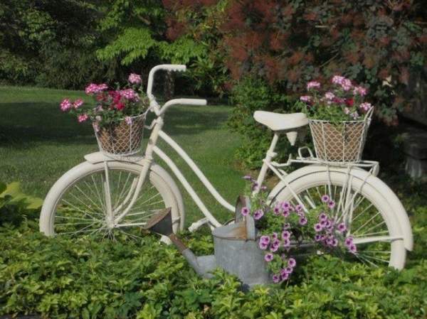 A white bicycle sits in a garden with flowers and a watering can.